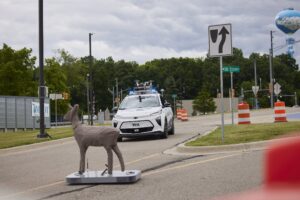 White car with sensors on the roof stops at intersection behind a deer dummy